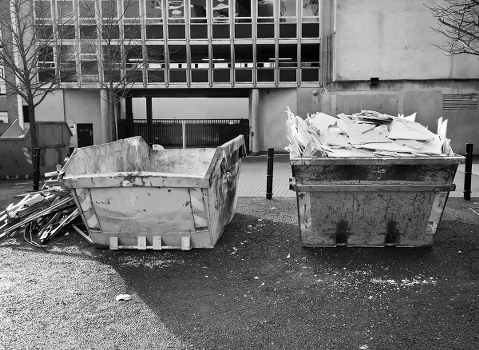 Two large industrial skips positioned side by side on a paved outdoor area, with the left skip partially filled with irregularly shaped concrete rubble and debris, showing rough, broken surfaces and a light gray color. The right skip is filled with a large pile of white or light-colored paper waste, including flattened cardboard and crumpled sheets, stacking unevenly with some edges protruding over the rim. In the background, a modern concrete building with rectangular windows and a metal gate can be seen, along with a tree on the right side of the image. Small scattered debris and gravel are visible around the skips on the ground, indicating an active waste collection or clearance site. The overall scene is in black and white, emphasizing the textures and industrial setting typical of rubbish removal services in Stratford, with Garden Clearance Stratford providing professional waste management solutions.