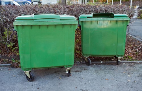 Two large green plastic wheelie bins with hinged lids are positioned side by side on a paved surface, with the larger bin on the left and the smaller one on the right. Both bins have visible wheels at the base for mobility, and the surface of the bins shows slight scuff marks and dirt, indicating regular use. The background features a hedge with brown and green foliage, suggesting an outdoor setting, possibly a residential driveway or street in Stratford. The lighting appears natural, likely from daylight, casting soft shadows on the ground. These waste containers are typical for rubbish collection services, as provided by companies such as Garden Clearance Stratford in the local area, aligning with the waste removal needs of households or businesses in the vicinity.