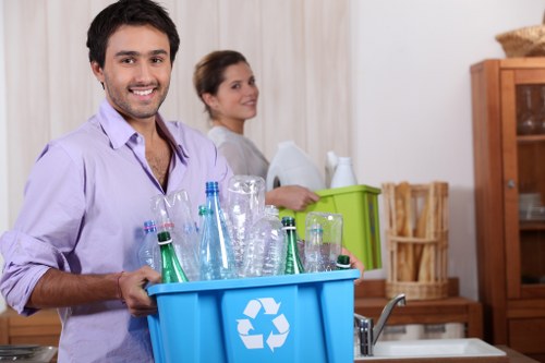 A man with dark hair, wearing a light purple shirt, is smiling and holding a blue recycling bin filled with various glass bottles and jars, placed on a kitchen countertop. Behind him, a woman with brown hair tied back is seated on a kitchen chair, looking towards the camera, with a neutral expression. The kitchen features a light-colored curtain, a wooden cabinet, and a green box or container on the work surface. The scene suggests a household effort to sort and prepare recyclable waste, such as glass bottles, possibly part of a rubbish removal process by Garden Clearance Stratford. The background includes typical kitchen elements like a sink and a wooden shelving unit. The overall environment appears well-lit, clean, and organized, reflecting domestic waste management activities that align with regional recycling standards and waste removal services in Stratford or nearby areas.
