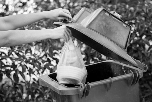 A person is seen disposing of waste into a large black and grey rubbish bin with an open lid, situated outdoors on a driveway or pavement area. The person's arms are visible, holding a plastic shopping bag filled with various items, including a white bowl and other unidentifiable objects. The lid of the bin is propped open, revealing an empty, dark interior. Surrounding the bin are bushes or hedges, indicating a garden or outdoor residential setting within Stratford or nearby. The scene reflects routine waste disposal activity relevant to rubbish removal services such as those offered by Garden Clearance Stratford, emphasizing the process of clearing household waste or garden debris, with natural lighting typical of daytime conditions. The environment suggests a tidy, well-maintained area, reinforcing the context of property and garden clearance solutions.