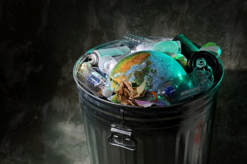 A close-up image of a silver metal rubbish bin filled with various types of waste, including plastic bottles, crumpled paper, and a small globe or spherical object with a map design on its surface. The bin is situated outdoors against a dark, slightly textured background, with the waste items piled high, some partially leaning over the edge. The scene highlights common household and environmental waste, emphasizing the importance of proper rubbish disposal and waste management services that Garden Clearance Stratford provides in Stratford and surrounding areas. The lighting focuses on the contents, creating reflections on the metallic surface of the bin and accents on the plastic bottles and the globe object, illustrating the variety of rubbish collected during clearance operations.