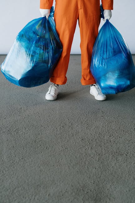 A person wearing orange overalls and white sneakers is standing on a gray concrete surface, holding two large blue plastic rubbish bags, one in each hand. The bags are semi-transparent, revealing cont