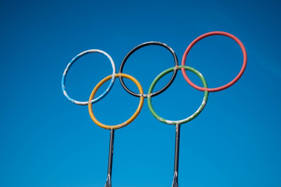 A close-up image of four metal hoops arranged to resemble the Olympic rings, each painted in vibrant colors — blue, black, yellow, green, and red — against a bright blue sky background. The hoops