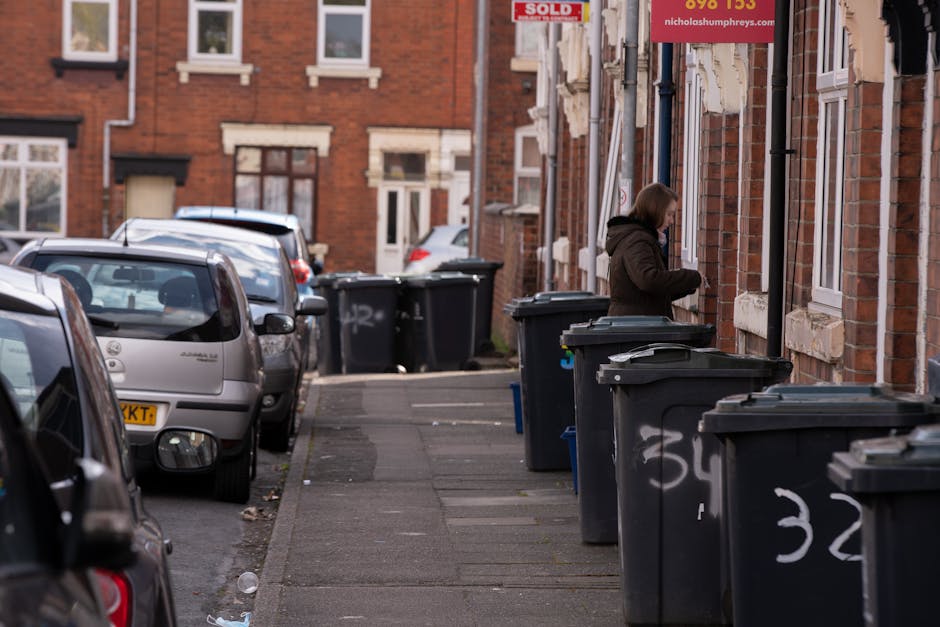 Five large wheeled rubbish bins with black plastic bodies and bright yellow lids are lined up in a row on a concrete pavement against a plain, light-colored wall. Each bin has a white oval label with 