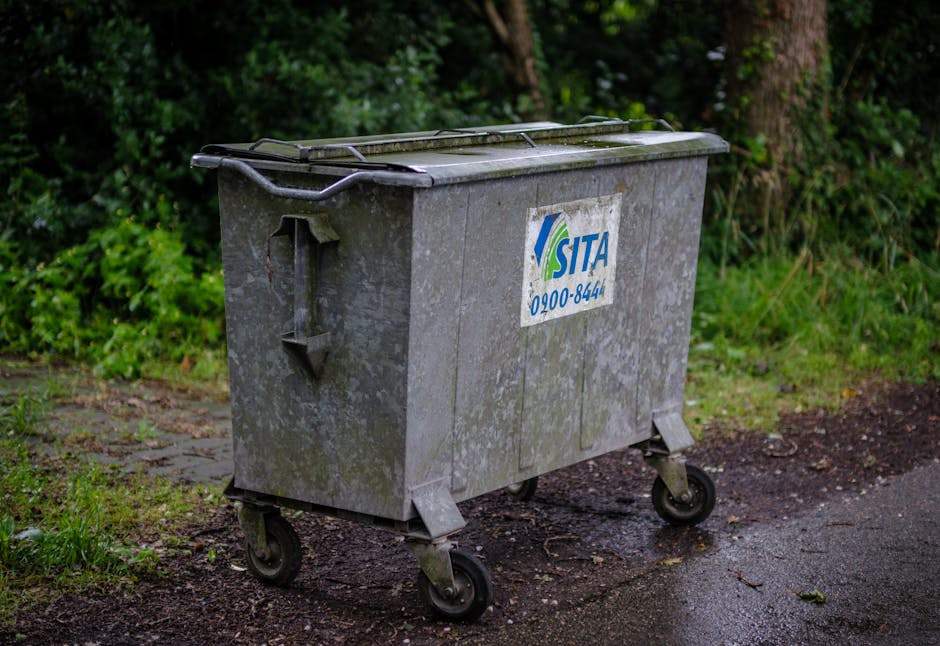 A white rubbish collection truck with an open rear hopper is positioned on a narrow cobblestone street, adjacent to a sidewalk lined with old, multi-storey buildings showing signs of weathering and pe